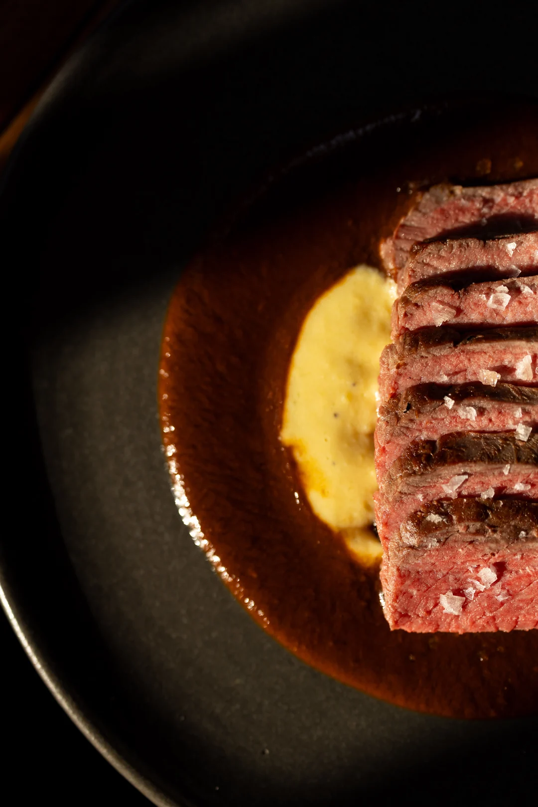 Detalle de carne en rebanadas con escamas de sal y salsa, servida en un plato oscuro